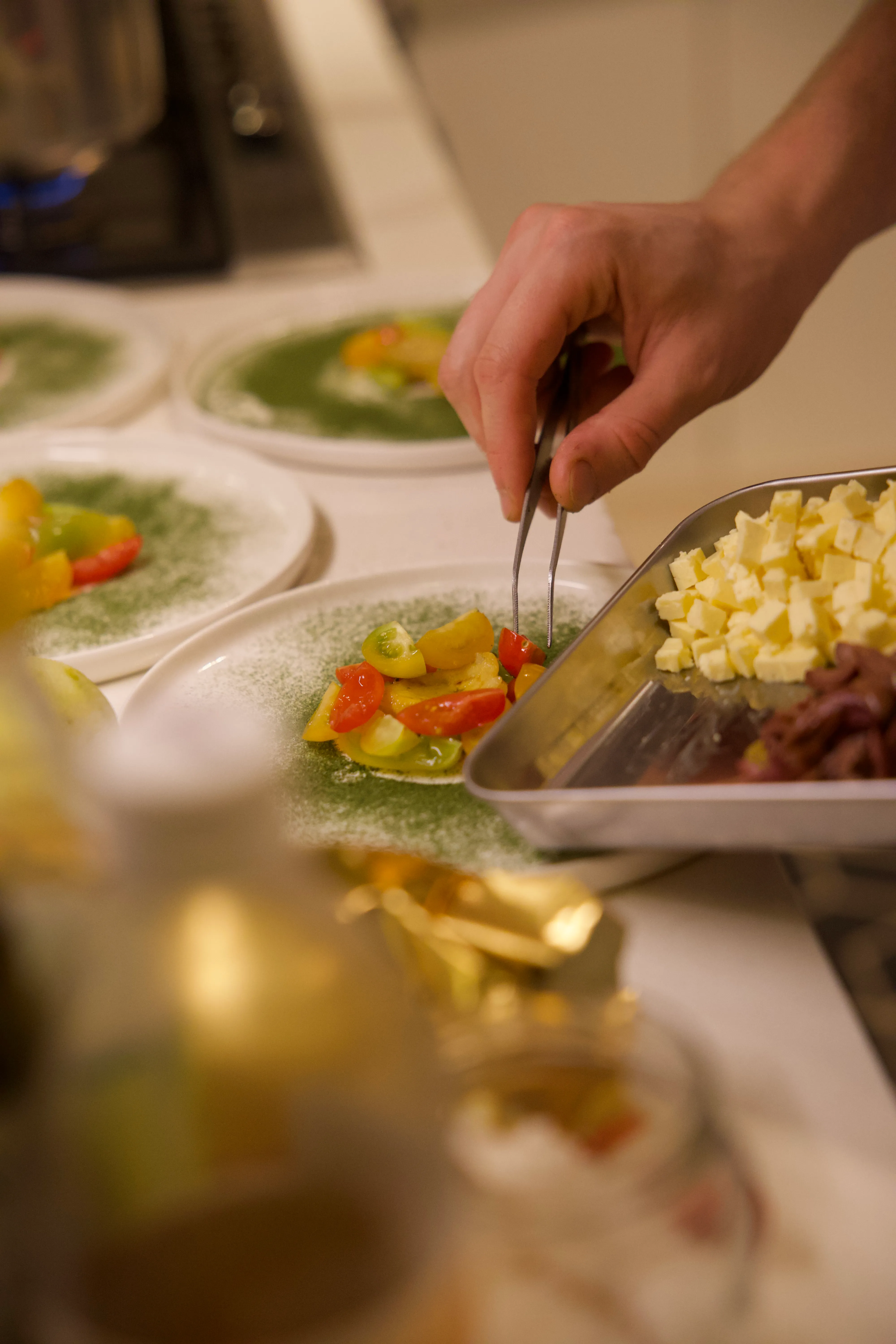 The chef’s hands dressing a plate with tweezers, tomatoes and feta laid out beside it.