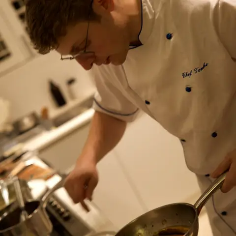 The chef, glasses on, spooning sauce over a plate of herb-crusted lamb.