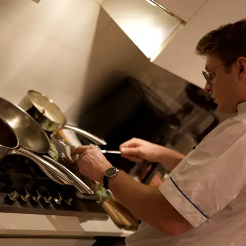 The chef tending pans at the stove, tasting a dish under the extractor light.