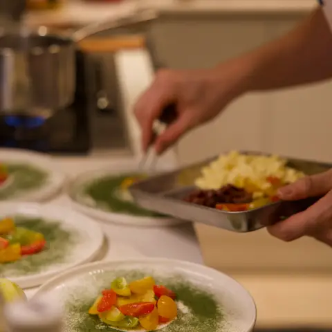 The chef’s hands, tray of feta and olives in one, tweezers plating tomatoes in the other.