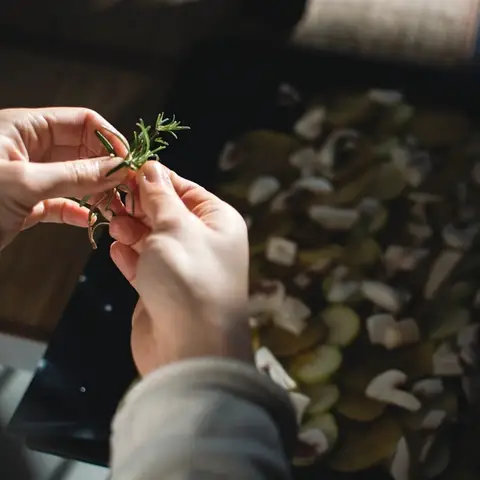 A hand laying a sprig of rosemary over a dark roasting tray of sliced mushrooms.