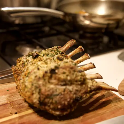 A whole herb-crusted rack of lamb resting on a wooden board beside the stove.