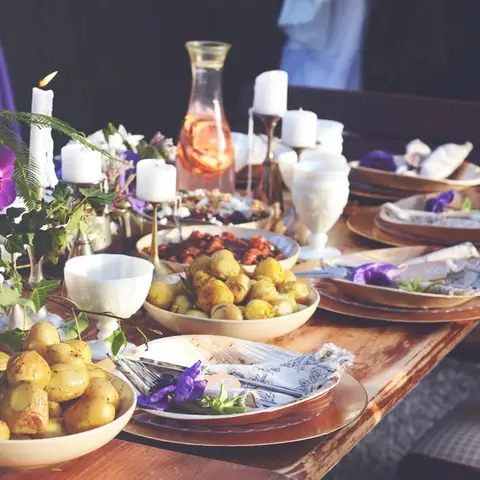 A long table laid for dinner by candlelight, with flowers, a carafe and bowls of new potatoes down its length.