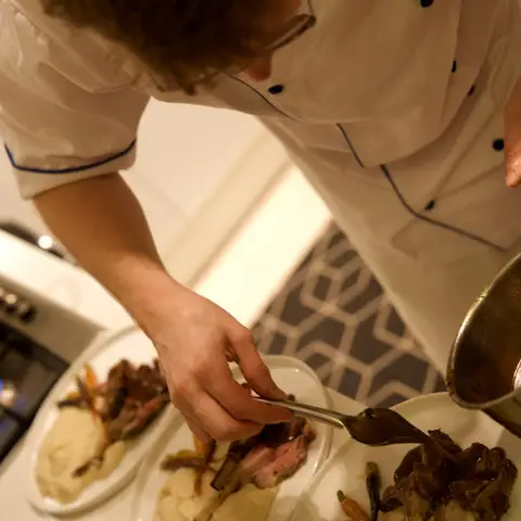 The chef spooning jus over plated lamb chops, parsnip purée and roasted baby carrots.