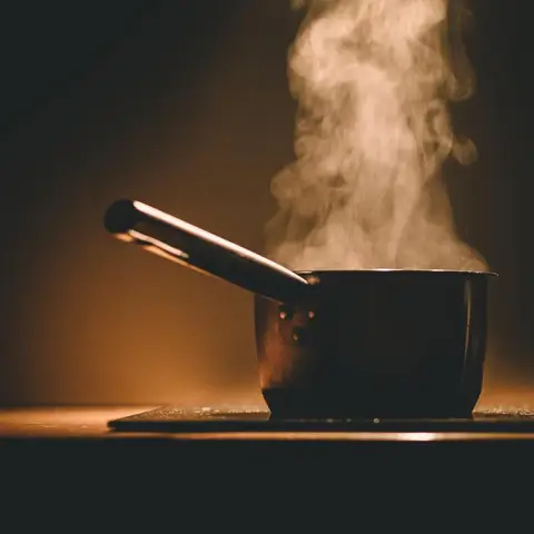 A saucepan steaming on a domestic hob, lit from one side by a low warm lamp.