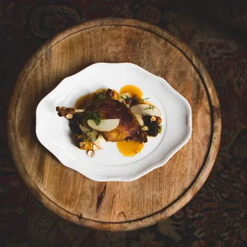 A single plated course of roast poultry with pear and a glossy sauce, on a white dish set on a round wooden board.