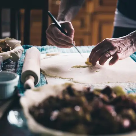 Hands rolling and trimming pastry on a kitchen table, a filled tart waiting in the foreground.