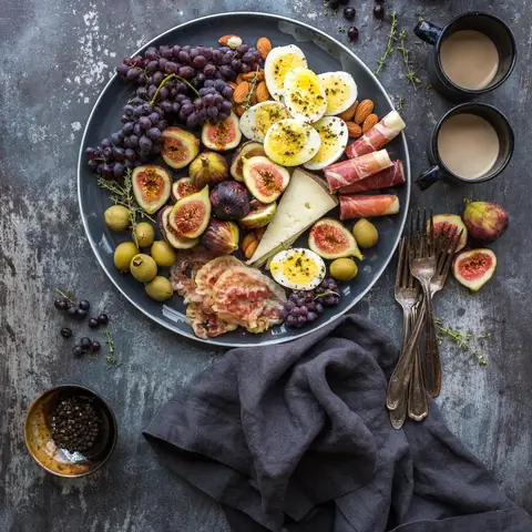 A sharing platter of figs, grapes, olives, cheese, cured meat and boiled eggs seen from above on a stone table, with two cups and a linen napkin.